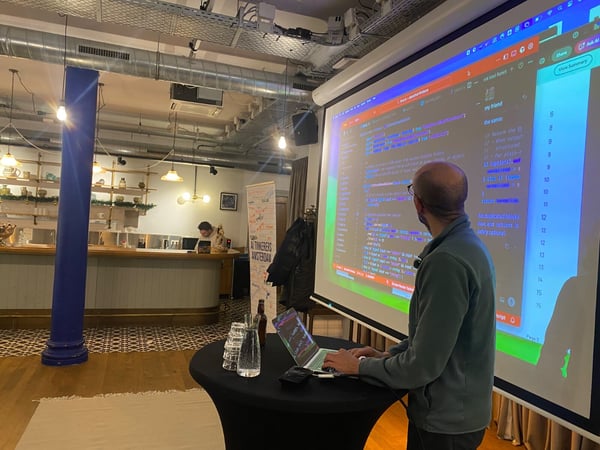A man is giving a technical presentation or code demonstration, standing in front of a large projection screen displaying syntax-highlighted code in an indoor event space with a bar area visible in the background.