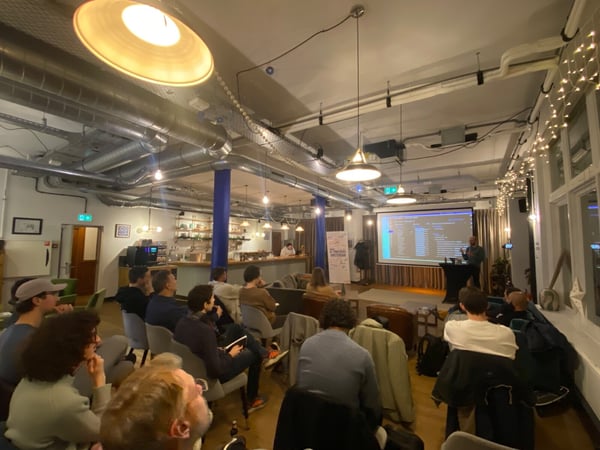 A candid photograph captures an indoor event, likely a tech meetup or presentation, where an audience is seated in a dimly lit, industrial-style venue, watching a presenter stand next to a screen displaying code or data.