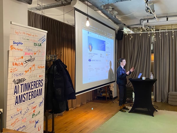 A photograph capturing an indoor tech event presentation, showing a speaker standing next to a small table while presenting content displayed on a large projection screen. To the left, a tall banner decorated with handwritten names and the title "AI TINKERERS AMSTERDAM" serves as a signature wall.