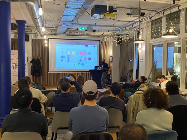 A photograph capturing an indoor technical presentation or meetup, showing a speaker standing next to a projection screen displaying a complex diagram, while an audience is seated facing the front.
