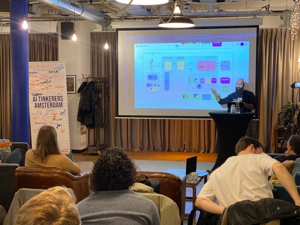 A photograph capturing a technical meetup or presentation event indoors, where a bearded presenter points to a projected screen displaying a complex system architecture diagram.