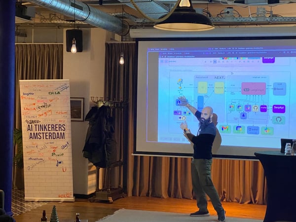 A man with a beard is giving a technical presentation, pointing at a complex system architecture diagram projected onto a large screen in a dimly lit indoor event space.