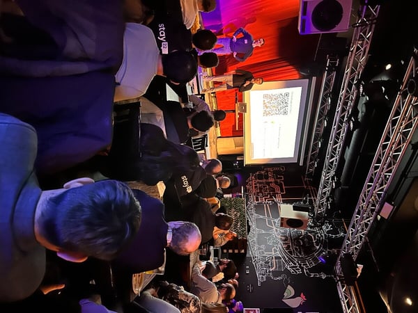 A photograph capturing a live presentation in an indoor venue, featuring a speaker on a stage with a red curtain backdrop and a seated audience.