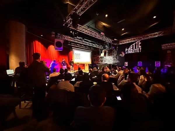 A wide-angle photograph of an indoor event where an audience is watching a presentation on a stage with red curtains and a projector screen.