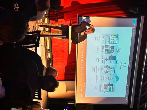 A man stands on a stage with a red curtain background, giving a presentation about QA CI/CD processes to an audience.
