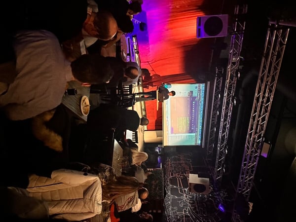 A person is delivering a presentation on a stage with a projector screen in a dimly lit venue, surrounded by an audience seated at tables.