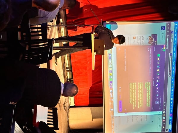 A person is giving a presentation on a stage with a large screen displaying a LinkedIn profile and a text generation tool, while audience members watch from the foreground.
