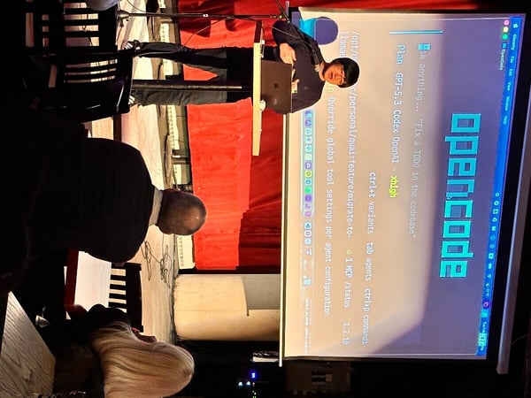 A man is giving a presentation on a stage in front of a projection screen displaying a software interface called 'opencode'. Two audience members are visible in the foreground, viewed from behind.
