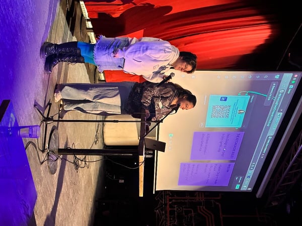Two women are on a stage giving a presentation, with one holding a microphone and the other standing behind a podium in front of a large projection screen.