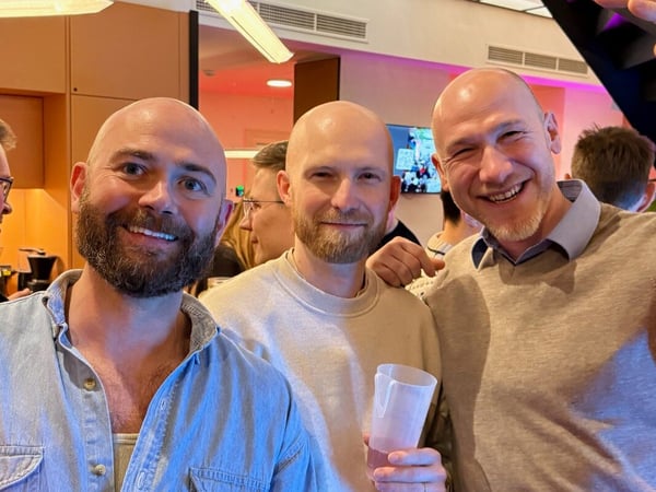 Three bald men with beards are smiling and posing for a photo during an indoor social event.