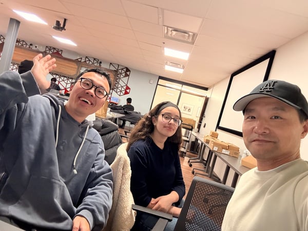 A group selfie captures three individuals—two men and one woman—smiling in an indoor office setting, which features a projection screen and geometric red and black wall decorations in the background.