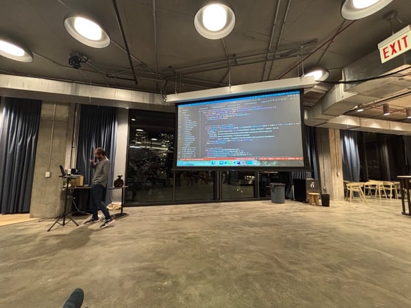 A wide-angle photograph captures a technical presentation taking place in an industrial-style event space at night, showing a presenter standing next to a laptop while code is displayed on a large projection screen.