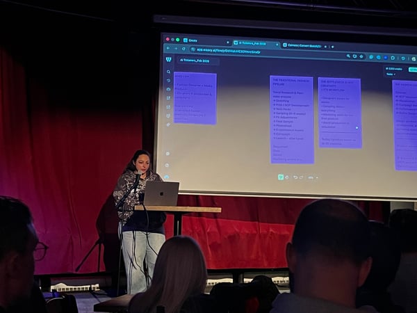 A woman is giving a presentation on a stage, standing behind a podium with a laptop in front of a large screen displaying a digital workspace.