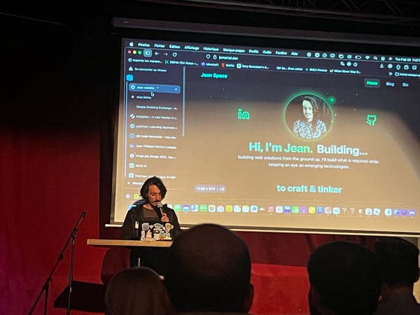 A person is giving a presentation on a stage, with a large screen behind them displaying a personal website and portfolio.