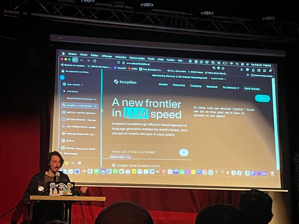 A person is giving a presentation in a dimly lit room, standing next to a large projector screen displaying a website for 'Inception' and its AI model capabilities.