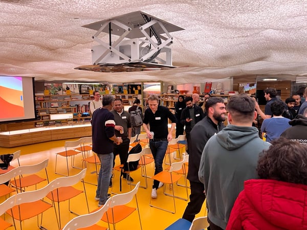 A candid photograph captures a large group of people mingling and networking in a brightly colored indoor event space featuring a yellow floor, rows of orange and white chairs, and a unique cloud-like ceiling structure.