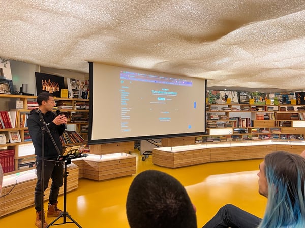 A man is giving a presentation, holding a microphone, in a uniquely designed indoor space featuring a bright yellow floor, curved wooden bookshelves lining the walls, and a highly textured, wavy ceiling.