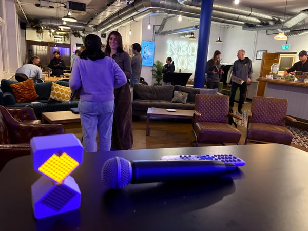 An indoor social space or lounge featuring people conversing in the background and a microphone with a glowing geometric light in the foreground.