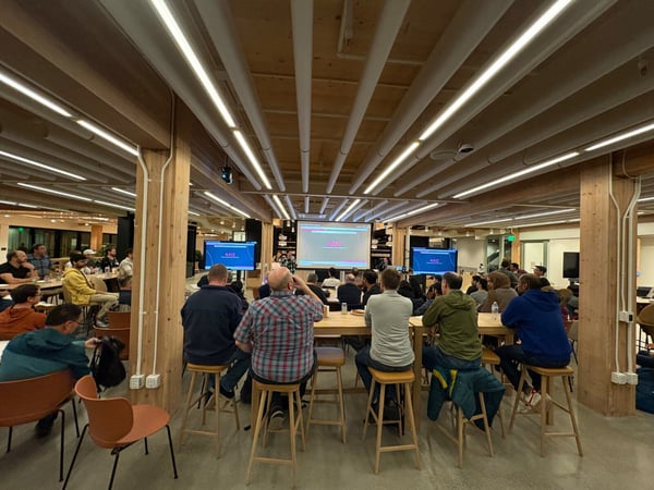 A photograph showing a large group of people gathered indoors at high tables, attending a presentation or meeting in a modern office space with exposed wood beams and linear ceiling lights.