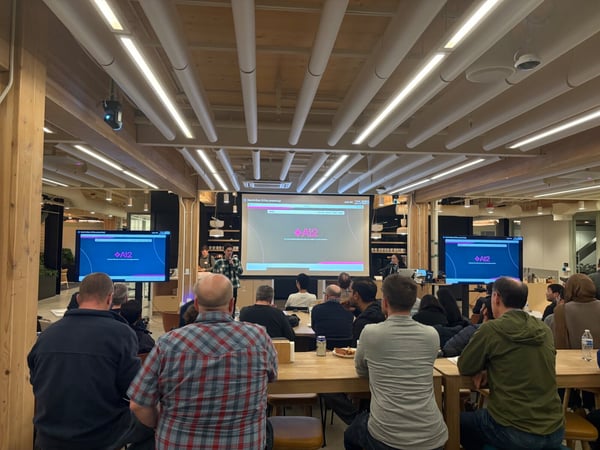 A photo capturing an indoor presentation or tech talk, showing a speaker addressing a seated audience in a modern office space characterized by wooden beams and linear lighting.