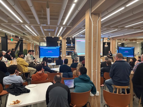 A wide-angle photograph capturing an indoor presentation or meeting taking place in a modern, open-plan office space, featuring a speaker addressing a large audience gathered around tables and chairs, with two large monitors displaying the 'AI2' logo.