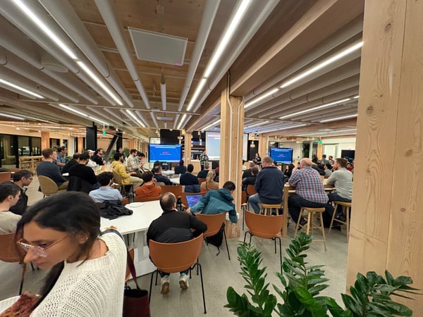 A candid photograph captures a large audience seated in a modern, wood-paneled office space attending a presentation, with two screens visible at the front of the room.