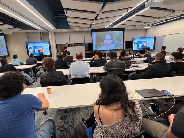 An indoor photograph of an audience seated at rows of desks in a modern lecture hall, watching a presentation displayed on multiple screens at the front of the room.