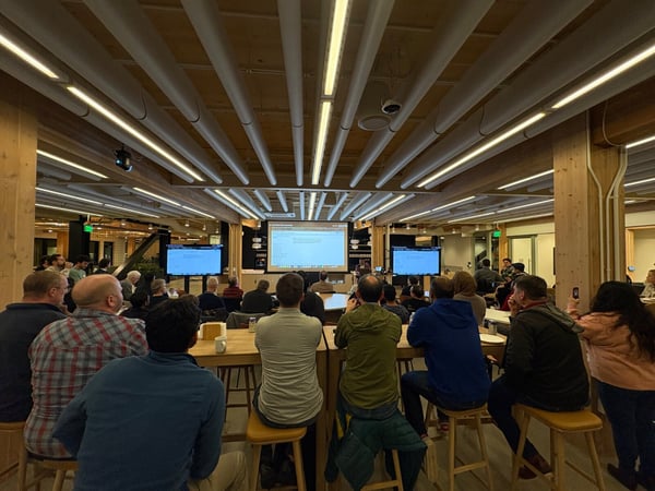 A photograph captures a large audience seated on high stools, facing forward to watch a presentation displayed on a central projected screen and two flanking monitors in a modern room featuring exposed wood beams and linear ceiling lighting.