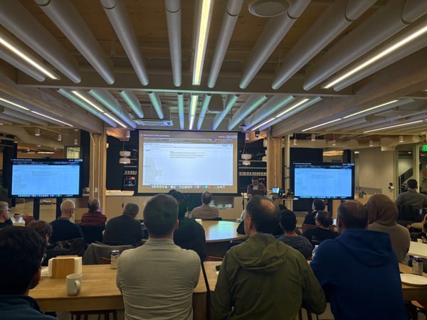 A photograph captures an indoor technical presentation where an audience is seated at wooden tables, facing a large central projected screen and two side monitors displaying technical data or code.