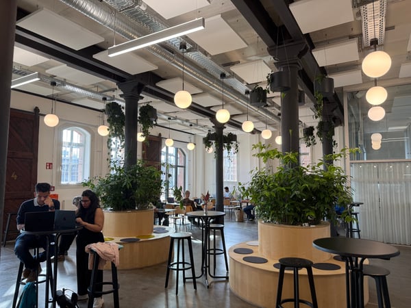 A modern, industrial-style coworking space or cafe featuring high ceilings, large pillars, hanging plants, and people working at tables with laptops.