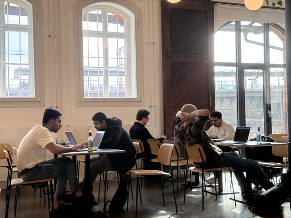A group of people are working on laptops at tables in a bright, high-ceilinged indoor space with large windows and wooden accents.
