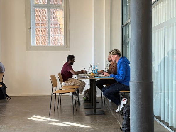Three young men are sitting at a long table in a bright, minimalist room, working on their laptops and talking.