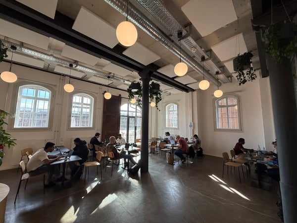 An indoor shot of a spacious, industrial-style co-working space or cafe where several people are working on their laptops at various tables.