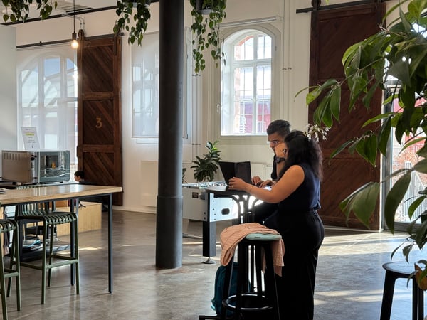 Two people are collaborating on a laptop at a high table in a spacious, modern office or cafe environment with industrial features.
