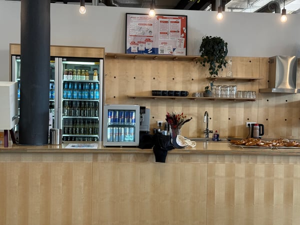A modern indoor bar or kitchen area featuring light-colored wood paneling, stocked drink refrigerators, and pastries on a counter.
