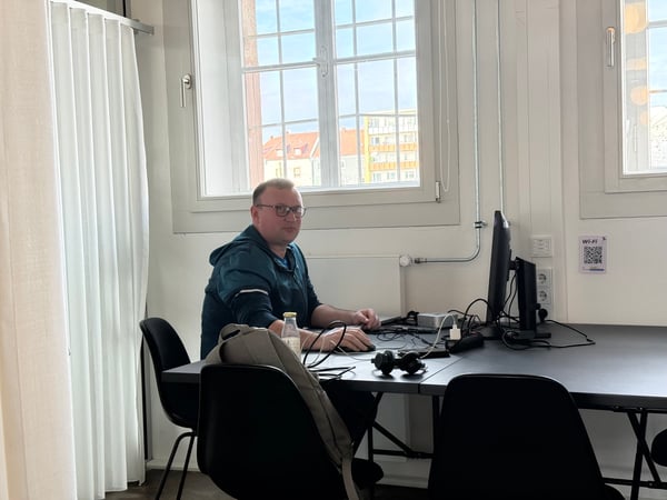 A man wearing glasses and a teal hoodie sits at a black desk with a computer in a bright office space, looking towards the camera.