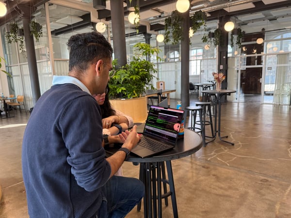 Two people are sitting at a high table in a modern, open-plan office space, focused on a laptop screen displaying lines of code.