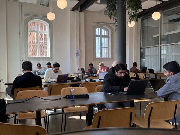 A group of people are working on their laptops in a bright, modern co-working space or office with high ceilings and large arched windows.