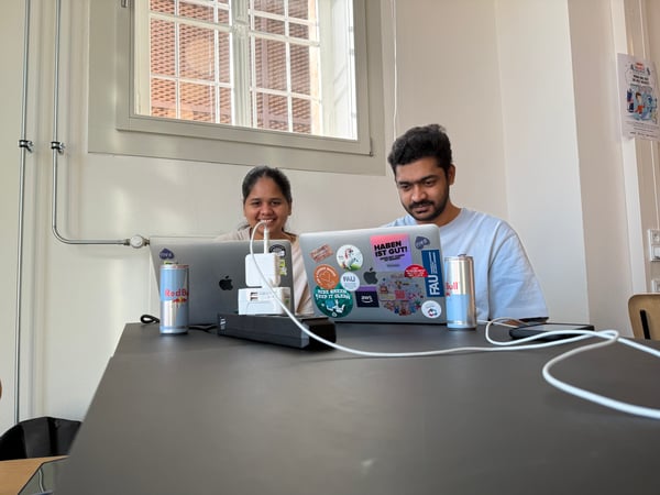 A man and a woman are sitting at a dark table, focused on their laptops which are covered in various stickers.