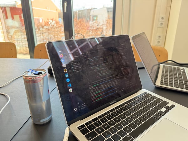 A close-up photograph of a MacBook Air displaying Python code in a text editor, positioned next to a silver energy drink can on a dark desk in an office setting.