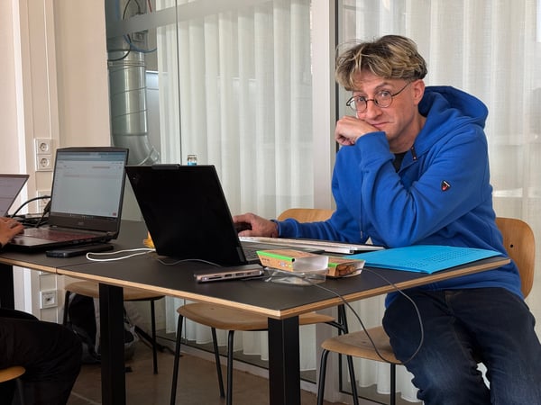 A man in a blue hoodie sits at a desk with two laptops, looking towards the camera with a thoughtful expression in an office environment.