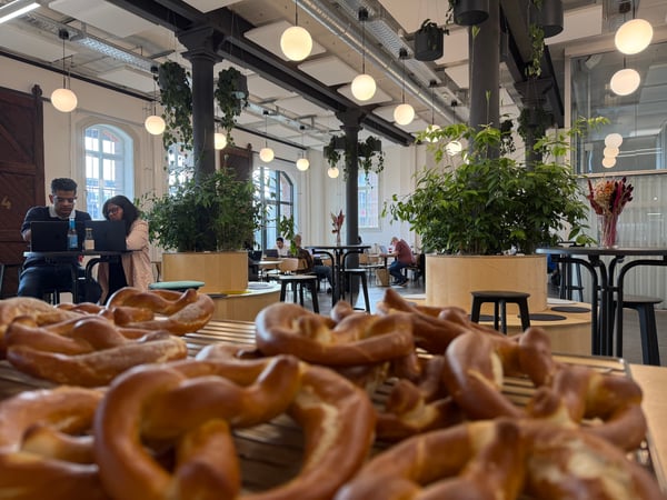 A wide-angle photograph of a modern indoor coworking space or cafe, featuring a tray of fresh pretzels in the foreground and people working on laptops in the background.