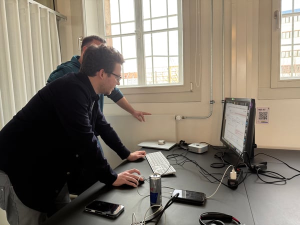 Two men are working at a desk with a computer monitor, keyboard, and mouse in an indoor office setting.