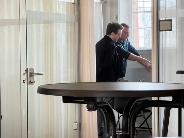 Two men are standing in a modern office environment, engaged in a discussion while looking at something out of view.