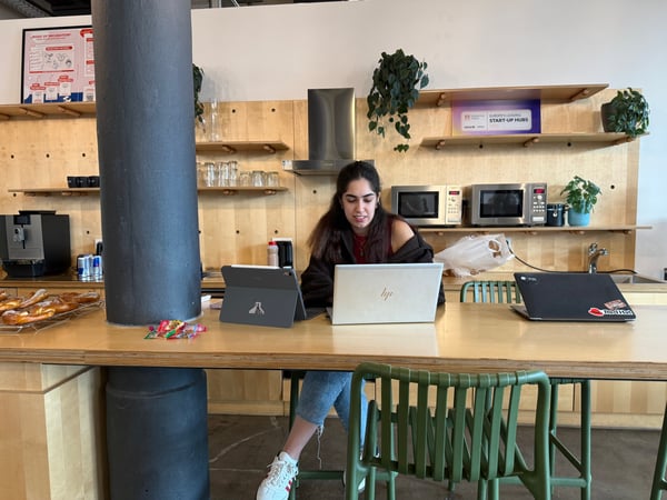 A woman sits at a long wooden table in a modern office kitchen area, working on a white laptop with other devices nearby.