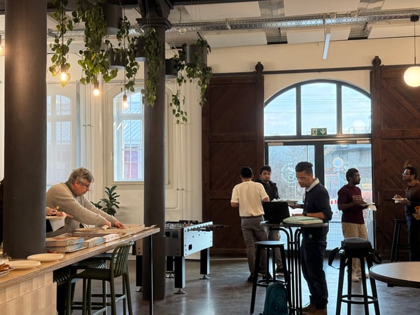 A group of people are gathered in a modern, high-ceilinged office social area with hanging plants and large wooden doors. One person is opening multiple pizza boxes on a long table while others stand nearby with plates.