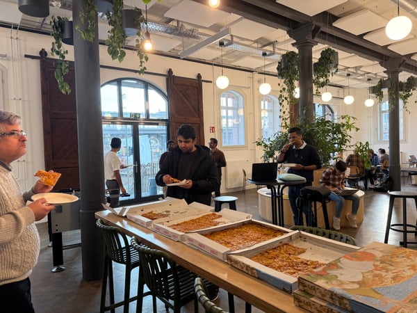 A group of people are gathered in a modern, open-plan office space, enjoying a pizza lunch from several large boxes laid out on a long wooden table.