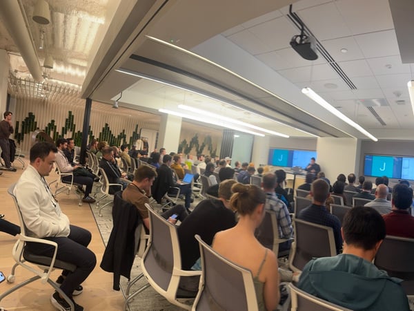 A photograph showing a large audience seated in a modern conference room, attending a presentation given by a speaker at the front. Two large screens behind the speaker display a bright blue background with the letter 'J'.