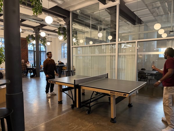Two people are playing table tennis in a spacious, modern office or common area with industrial-style architecture and hanging plants.