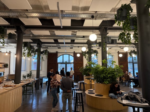 A wide-angle photograph of a modern, industrial-style communal space or office featuring high ceilings, hanging plants, and people socializing.
